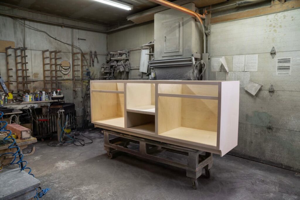 Unfinished plywood cabinet carcass on a rolling cart in the Rissler woodshop, with hand tools and parts racks lining the walls behind it.