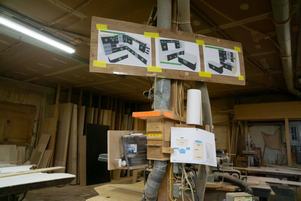 Printed kitchen design plans taped to a board in the Rissler woodshop, posted above the machinery for the workers to follow.