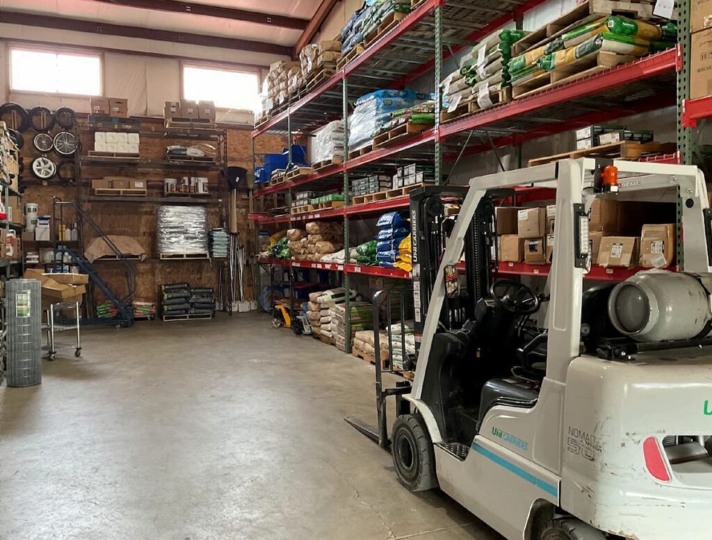Forklift beside pallet racking stocked with bagged goods and building supplies inside the Shady Lane Hardware warehouse.