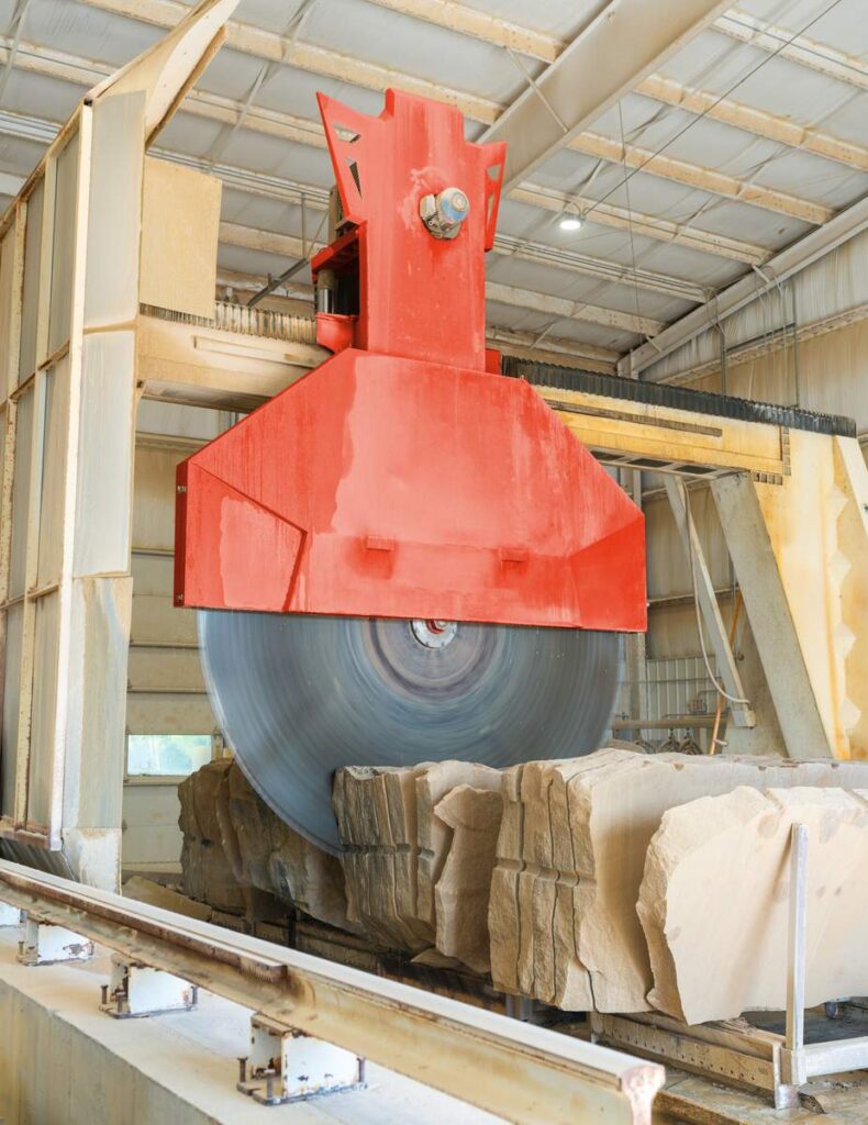 A red-housed circular saw with a diamond-tipped blade nearly 12 feet across cuts into a row of sandstone blocks inside the mill.