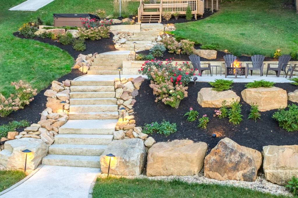 A landscaped hillside at dusk with sandstone stair treads, boulder retaining walls, and a lit stone patio with a fire pit.