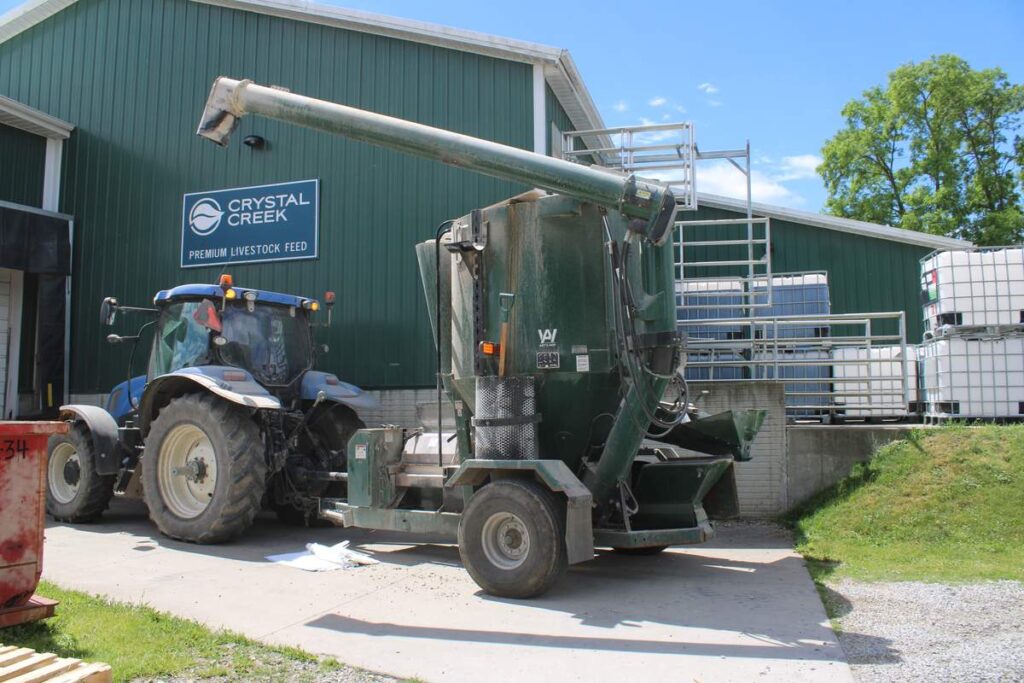 A blue tractor hitched to a green mobile feed grinder-mixer parked outside a warehouse with a Crystal Creek Premium Livestock Feed sign.
